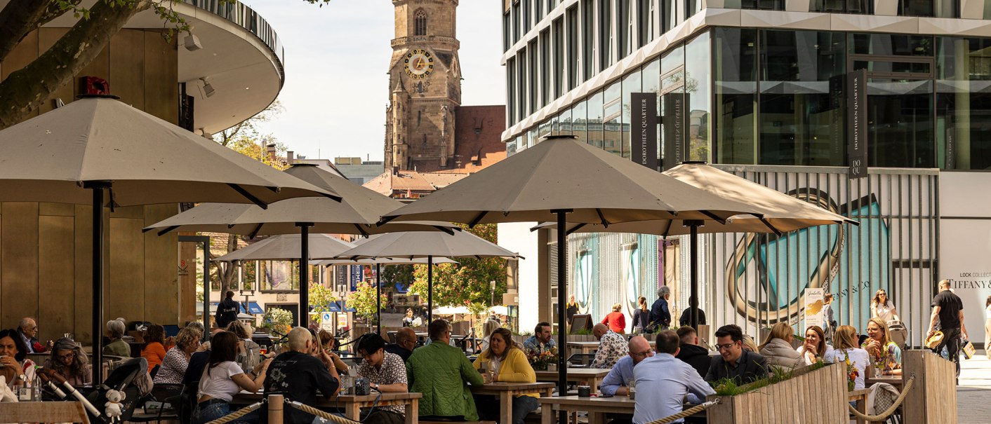 Menschen genie&szlig;en das sonnige Wetter im Dorotheenquartier in Stuttgart. Sie sitzen unter Sonnenschirmen, w&auml;hrend im Hintergrund eine Kirche zu sehen ist., &copy; Stuttgart-Marketing GmbH, Sarah Schmid