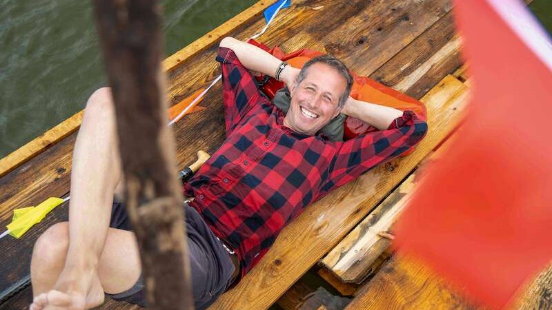 Ein Mann in einem karierten Hemd liegt entspannt auf einem Holzfloß auf dem Wasser. Er lächelt und hat die Arme hinter dem Kopf verschränkt., © Theaterhaus Stuttgart e.V. Ein Mann in einem karierten Hemd liegt entspannt auf einem Holzfloß auf dem Wasser. Er lächelt und hat die Arme hinter dem Kopf verschränkt., © Theaterhaus Stuttgart e.V.