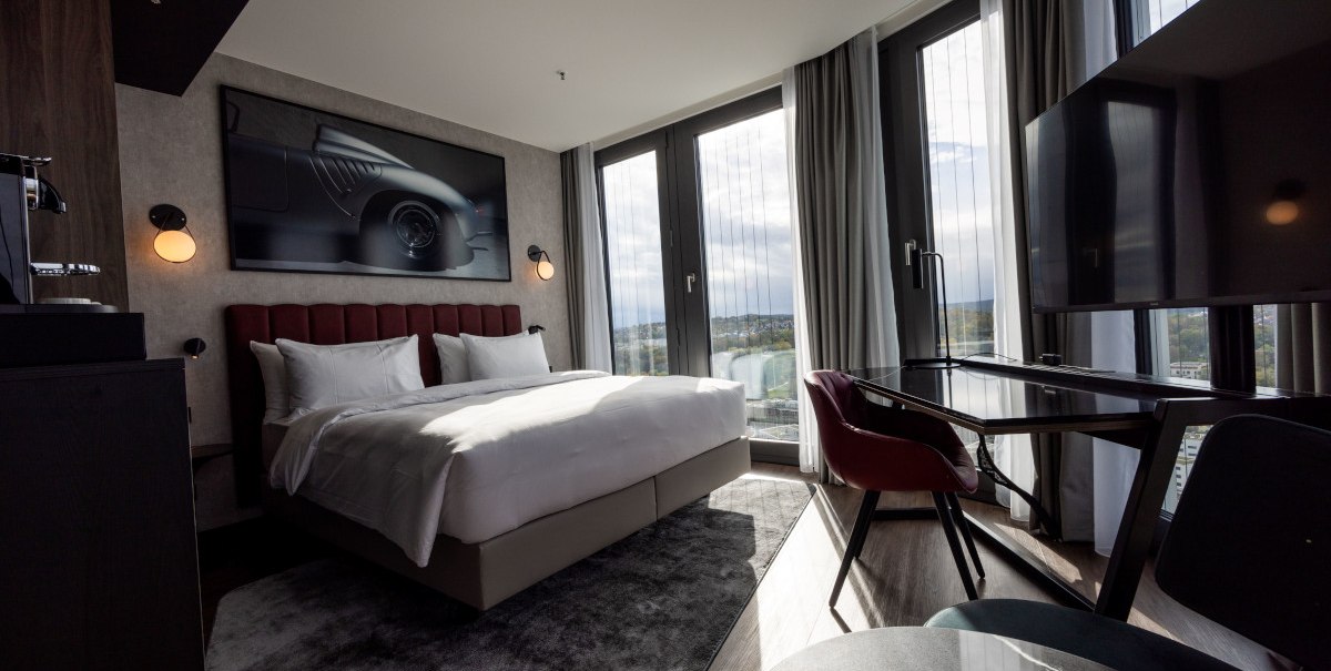 Modernes Eckzimmer mit großem Bett, Schreibtisch, Panoramafenstern und stilvollem Dekor. Ein Bild eines Autos hängt an der Wand., © Radisson Hotel Group Modernes Eckzimmer mit großem Bett, Schreibtisch, Panoramafenstern und stilvollem Dekor. Ein Bild eines Autos hängt an der Wand., © Radisson Hotel Group