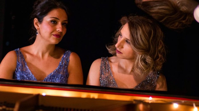 Zwei Frauen in glitzernden Kleidern sitzen nebeneinander an einem Flügel und schauen sich an. Der Hintergrund ist dunkel gehalten., © Blanc&Noir Zwei Frauen in glitzernden Kleidern sitzen nebeneinander an einem Flügel und schauen sich an. Der Hintergrund ist dunkel gehalten., © Blanc&Noir