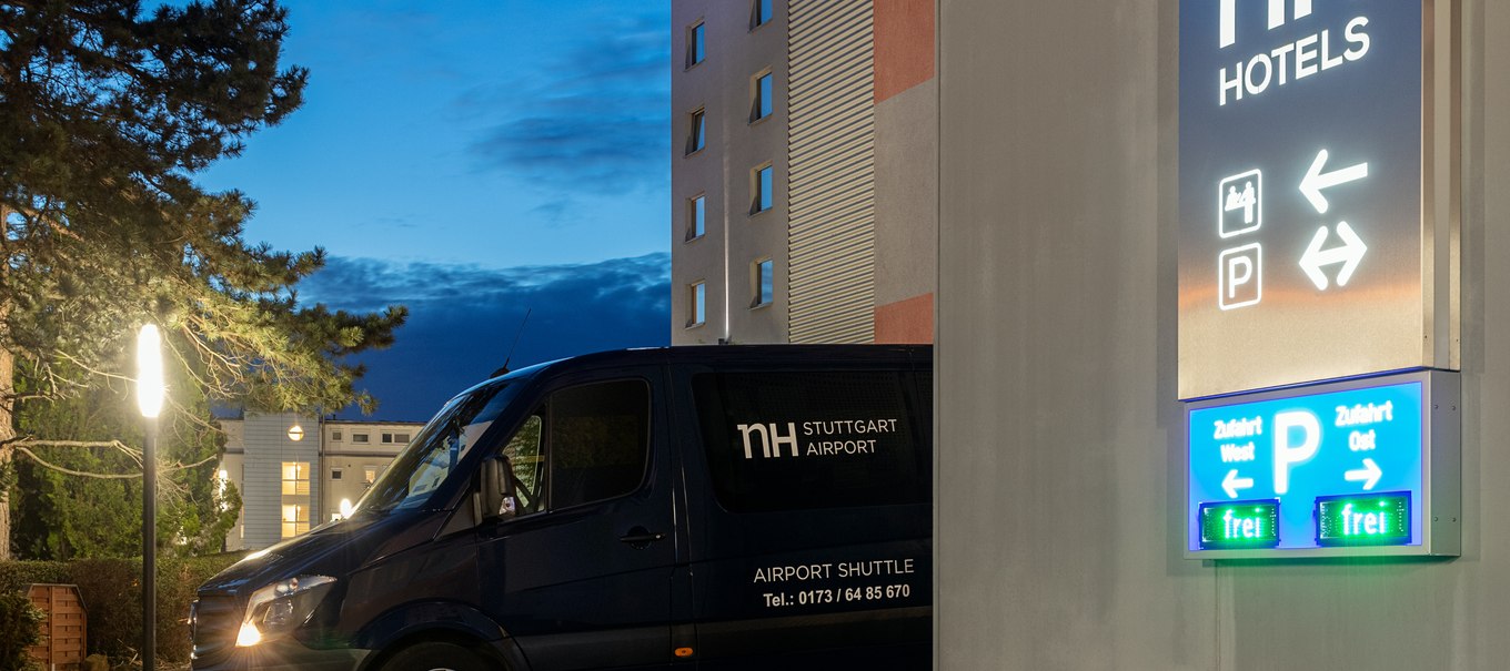 Ein Shuttlebus des NH Hotels Stuttgart Airport steht vor dem beleuchteten Hotelgebäude bei Dämmerung. Ein Schild weist auf Parkmöglichkeiten hin., © NH Hotel Stuttgart Airport