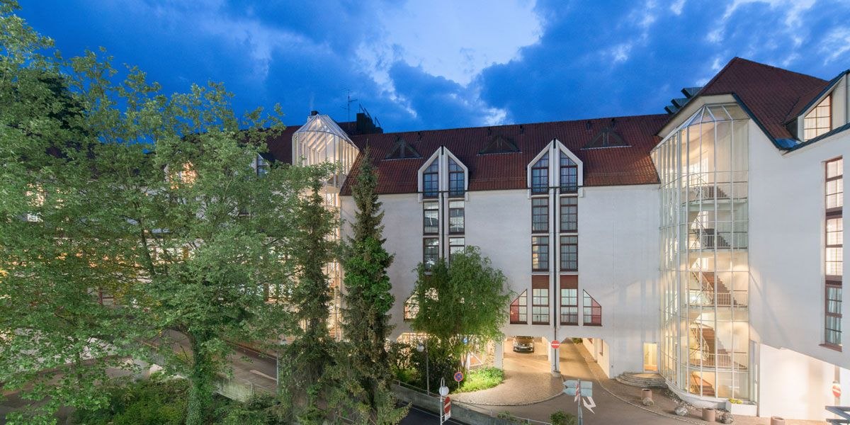 Das Best Western Plus Hotel am Schlossberg bei Dämmerung, mit beleuchteten Fenstern und einem gläsernen Treppenhaus, umgeben von Bäumen., © Best Western Plus Hotel am Schlossberg