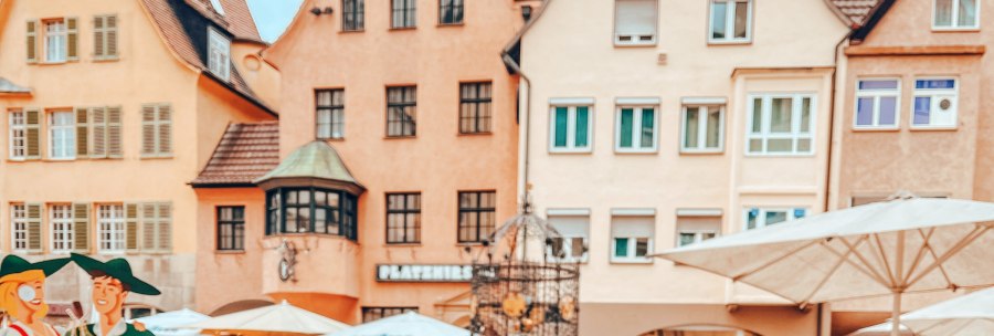 The Hans im Gl&uuml;ck fountain in front of historic buildings. Parasols and figures can be seen in the foreground. The houses have red roofs and many windows., &copy; SMG, takemyhearteverywhere