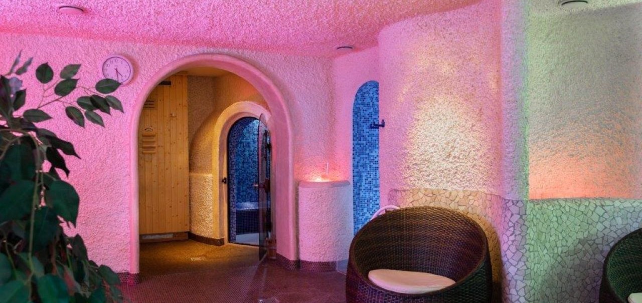 Ein Spa-Bereich mit bunten Lichtern, Rattan-Sesseln und einem Dampfbad-Eingang. Die Wände sind mit Mosaikfliesen verziert., © Kronen Hotel GmbH Ein Spa-Bereich mit bunten Lichtern, Rattan-Sesseln und einem Dampfbad-Eingang. Die Wände sind mit Mosaikfliesen verziert., © Kronen Hotel GmbH