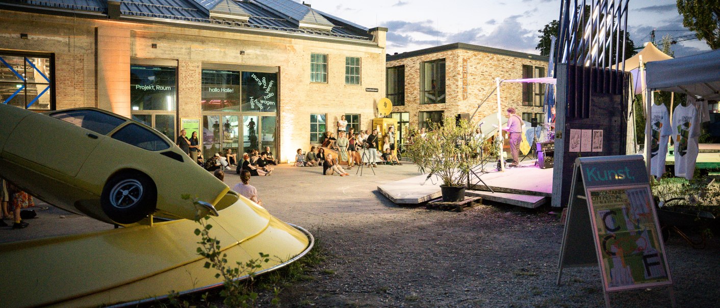 Evening event at the Kunstverein Wagenhalle with an art installation of a yellow car and people sitting in the square watching a performance., © Atelier Oradoro