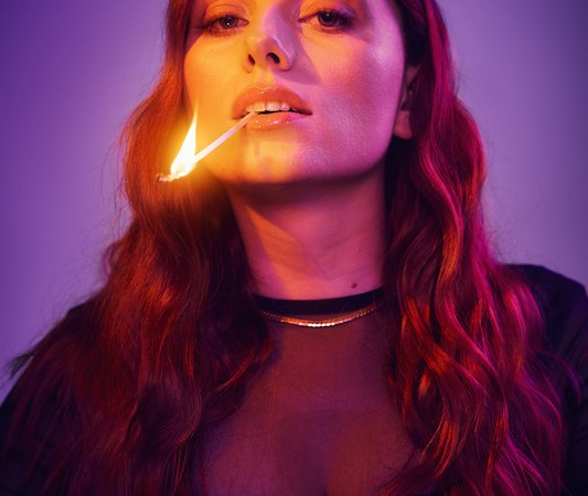 Woman with long, wavy hair holds a burning match in her mouth. She is wearing a black top and is bathed in violet light., © Rosenau Kultur e.V. Woman with long, wavy hair holds a burning match in her mouth. She is wearing a black top and is bathed in violet light., © Rosenau Kultur e.V.