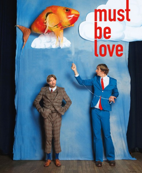 Zwei M&auml;nner in Anz&uuml;gen posieren vor einem blauen Hintergrund mit Wolken und einem fliegenden Fisch. Der Text 'must be love' ist sichtbar., &copy; Theaterhaus Stuttgart e.V.