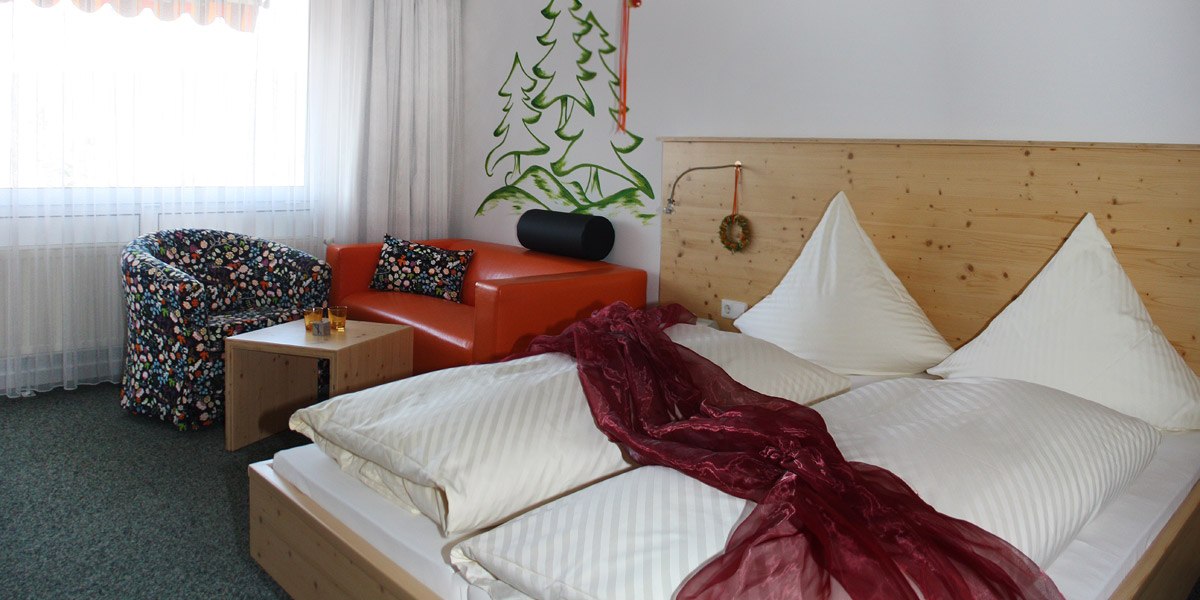 Double room with wooden bed, red sofa, patterned armchair and wall painting of trees. A red scarf lies on the bed., &copy; Hotel Hochwald