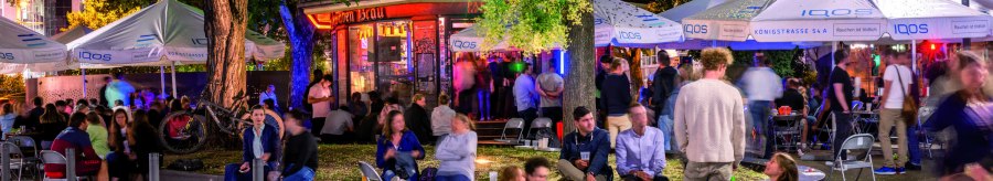 Lively outdoor area with people under illuminated umbrellas and trees. Some are sitting on chairs, others on the ground. Colorful lights in the background., © TMBW, Gregor Lengler