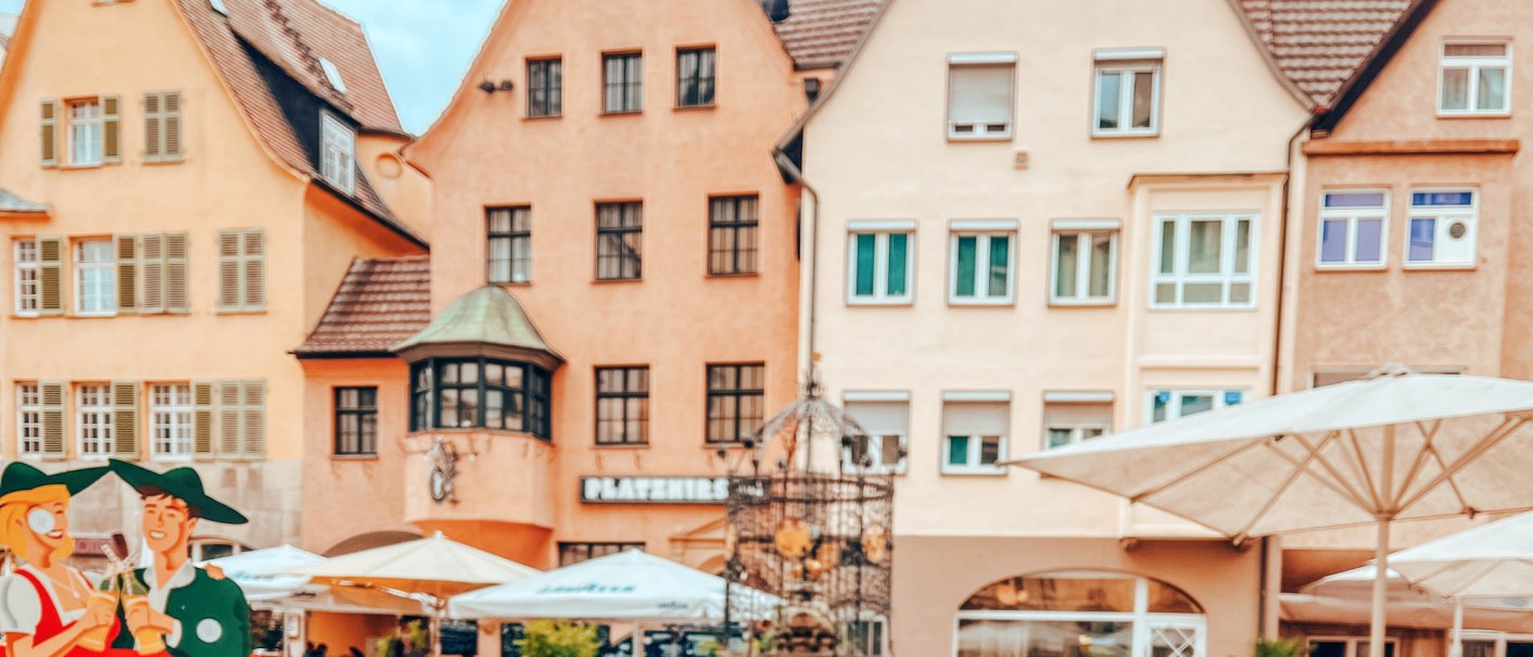 Der Hans-im-Glück-Brunnen vor historischen Gebäuden. Im Vordergrund sind Sonnenschirme und Figuren zu sehen. Die Häuser haben rote Dächer und viele Fenster., © SMG, takemyhearteverywhere