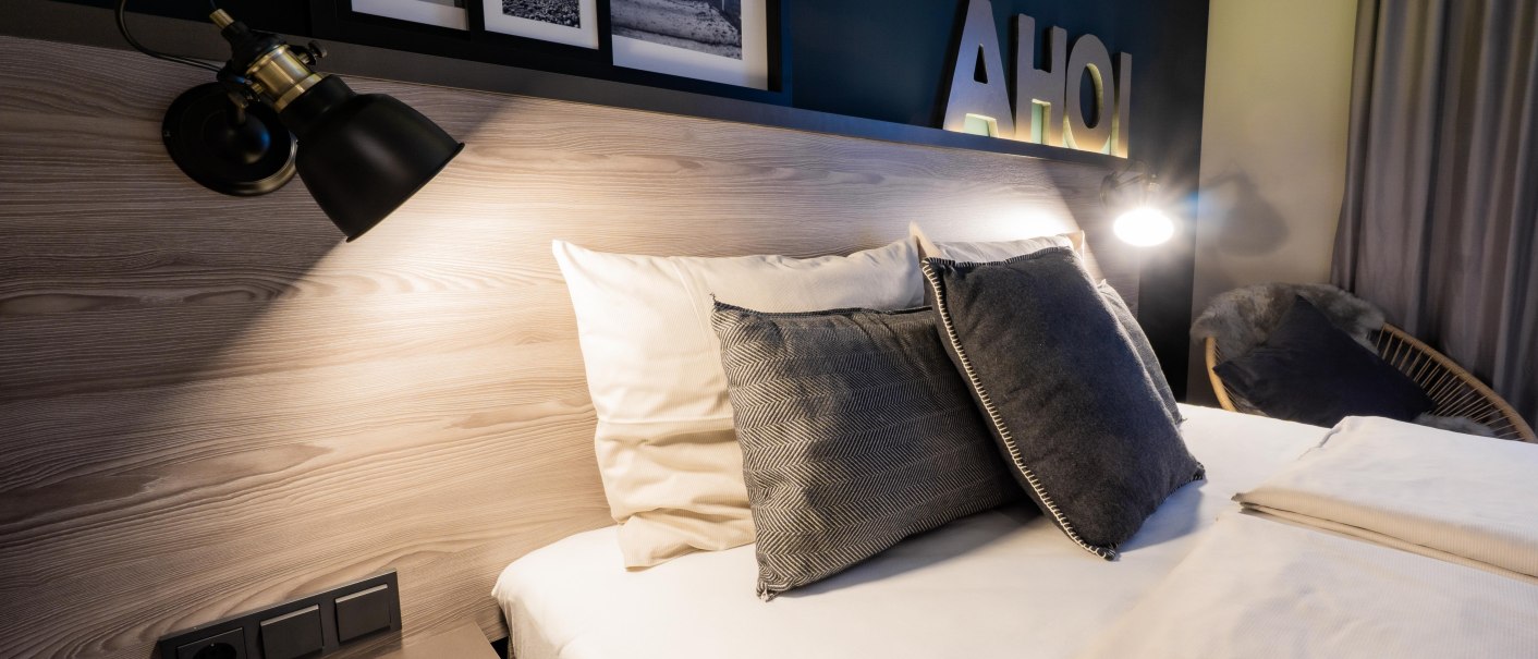 Modernes Schlafzimmer mit einem Bett, grauen Kissen, Wandlampen und Dekorationen wie Bilderrahmen und dem Schriftzug 'AHOI' an der Wand., &copy; HARBR. hotel Ludwigsburg / DQuadrat Living GmbH