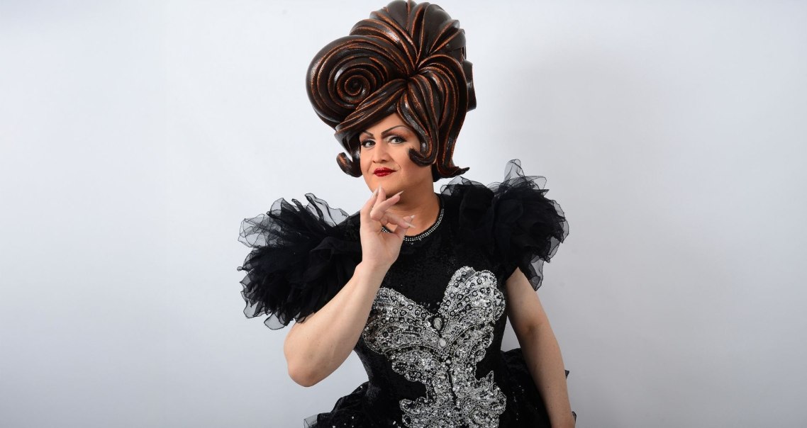 Person in auffälligem Kostüm mit großer Perücke und glitzerndem Kleid posiert vor weißem Hintergrund., © Friedrichsbau Varieté Person in auffälligem Kostüm mit großer Perücke und glitzerndem Kleid posiert vor weißem Hintergrund., © Friedrichsbau Varieté