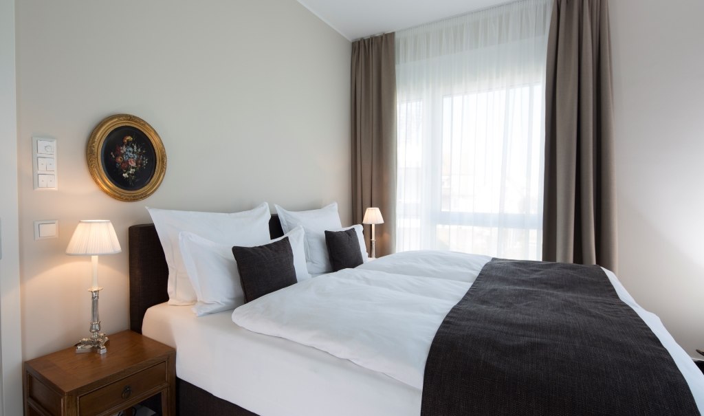 Elegant bedroom with large bed, white and dark cushions, bedside lamps and a round painting on the wall., © Hotel LOGIS Elegant bedroom with large bed, white and dark cushions, bedside lamps and a round painting on the wall., © Hotel LOGIS