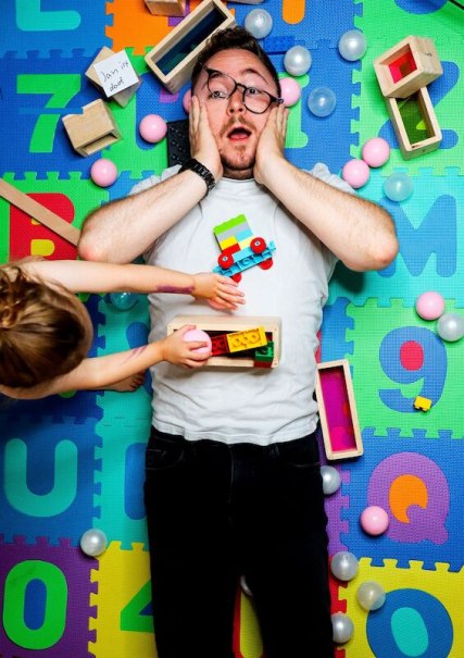 Surprised man lying on a colorful play mat, surrounded by building blocks and toys. A child hands him a toy. Humorous scene., &copy; Theaterhaus Stuttgart e.V.