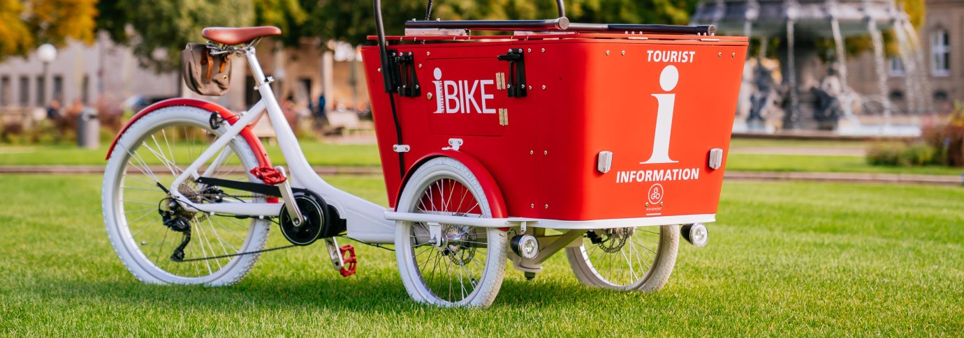Ein rotes Lastenfahrrad mit der Aufschrift 'I-Bike' steht auf einer Wiese vor einem Brunnen und einem Geb&auml;ude mit Kuppel., &copy; Stuttgart-Marketing GmbH, Thomas Niederm&uuml;ller