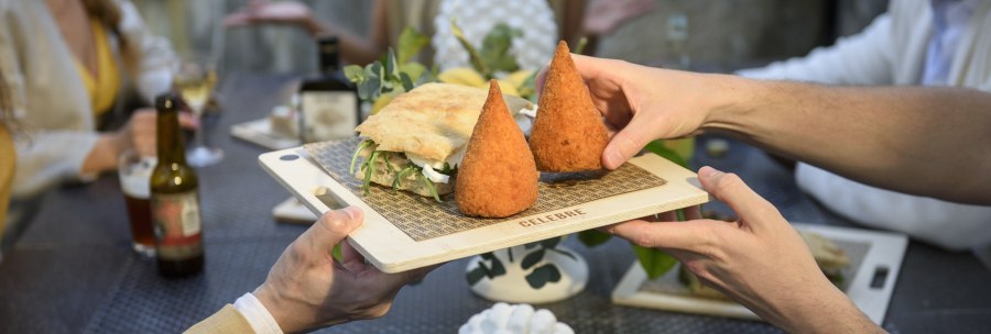 Two people pass a wooden board with arancini and a sandwich at a table with several guests., © CELEBRE Two people pass a wooden board with arancini and a sandwich at a table with several guests., © CELEBRE