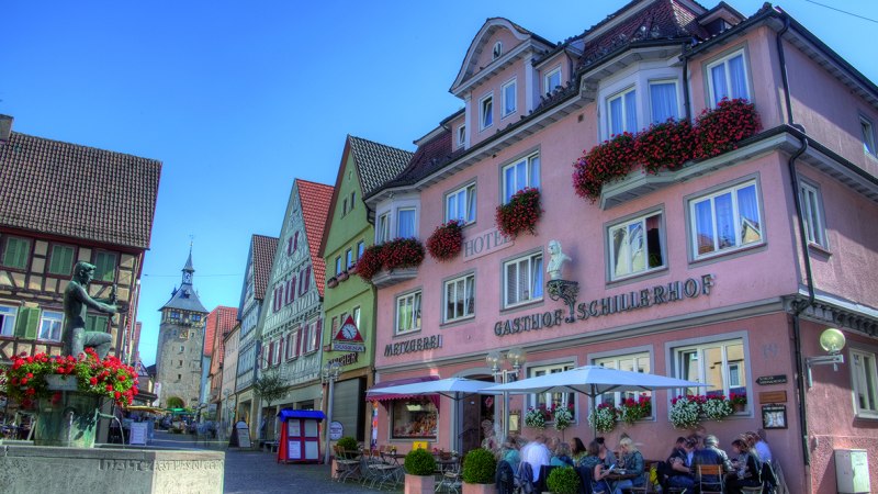 Hotel Schillerhof in einer malerischen Altstadt mit Fachwerkhäusern, einem Brunnen und Menschen, die im Freien sitzen. Sonniges Wetter und blauer Himmel., © Hotel Schillerhof