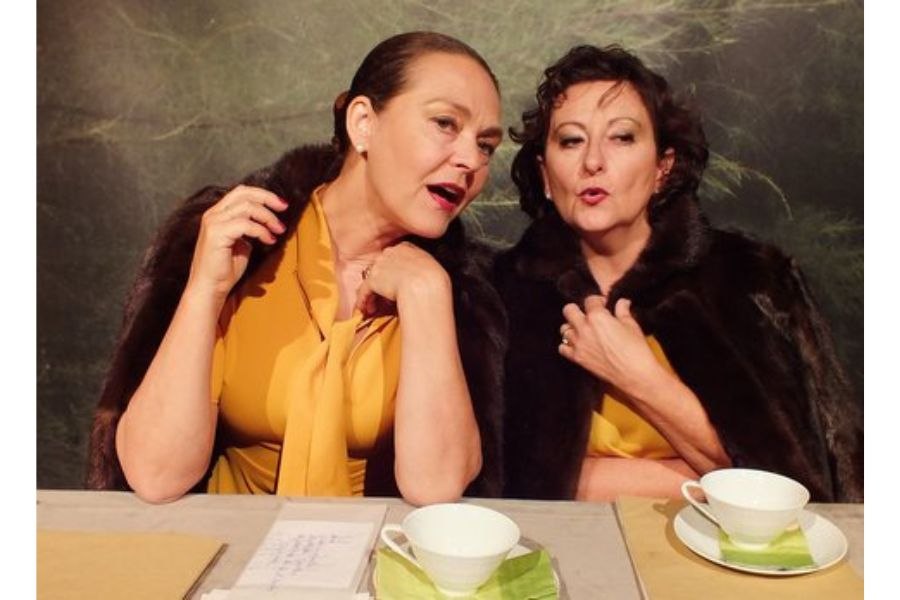 Zwei Frauen in gelber Kleidung und Pelzstolen sitzen an einem Tisch mit Teetassen und scheinen zu flüstern., © Fotocredit Theater La Lune Stuttgart