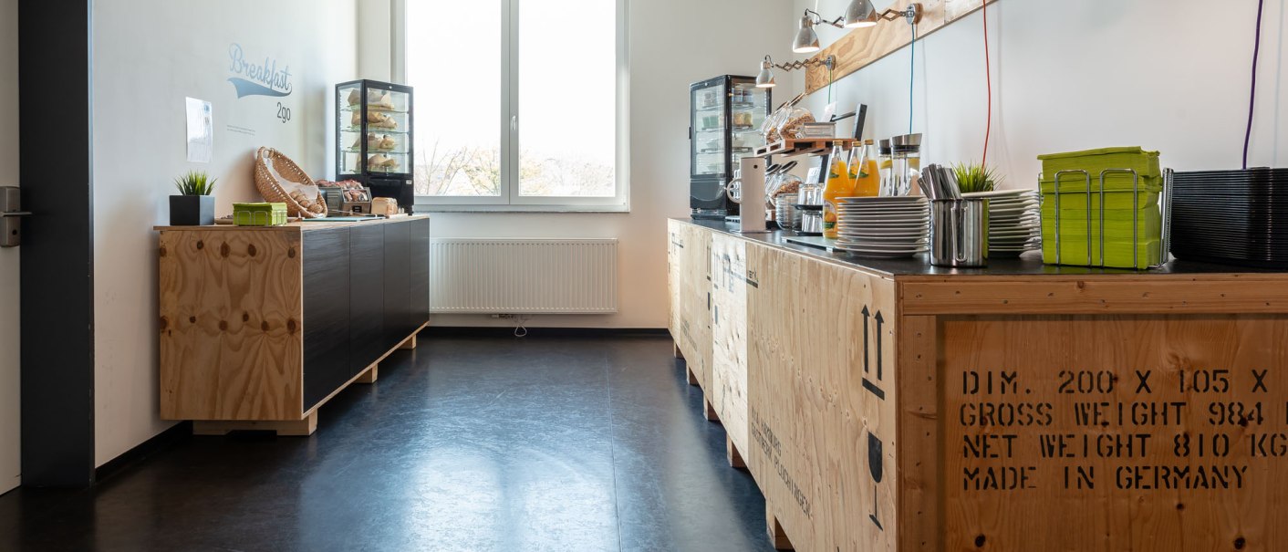 Moderner Frühstücksbereich mit Holztheken, Saftspendern und Gebäck in einem hellen Raum mit großem Fenster., © a2 HOTELS Plochingen Moderner Frühstücksbereich mit Holztheken, Saftspendern und Gebäck in einem hellen Raum mit großem Fenster., © a2 HOTELS Plochingen