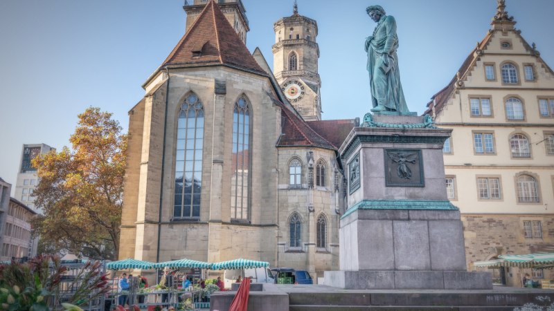Der Schillerplatz in Stuttgart zeigt eine Statue, historische Gebäude und Marktstände. Im Hintergrund ist eine Kirche mit Turm zu sehen., © SMG Martina Denker Der Schillerplatz in Stuttgart zeigt eine Statue, historische Gebäude und Marktstände. Im Hintergrund ist eine Kirche mit Turm zu sehen., © SMG Martina Denker