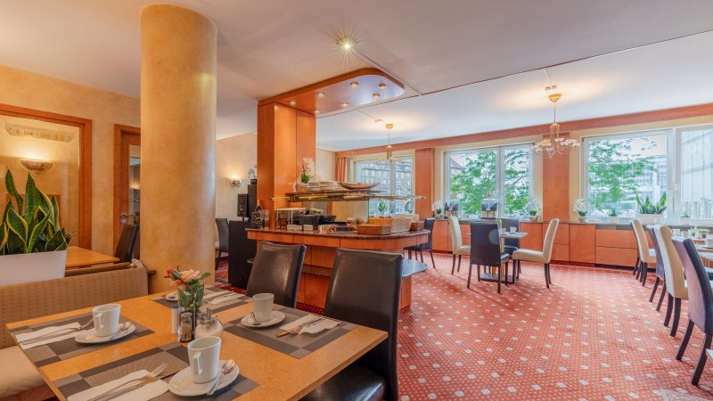 Eleganter Frühstücksraum mit gedeckten Tischen, Pflanzen und großen Fenstern. Warme Farben und gemütliche Atmosphäre., © PLAZA INN Rieker Stuttgart Hauptbahnhof