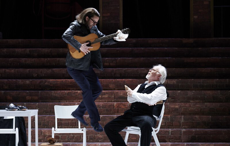 A man jumps with a guitar in his hand while another sits on a chair and watches. The scene takes place on a stage with a brick wall., © Württembergische Staatstheater Stuttgart A man jumps with a guitar in his hand while another sits on a chair and watches. The scene takes place on a stage with a brick wall., © Württembergische Staatstheater Stuttgart
