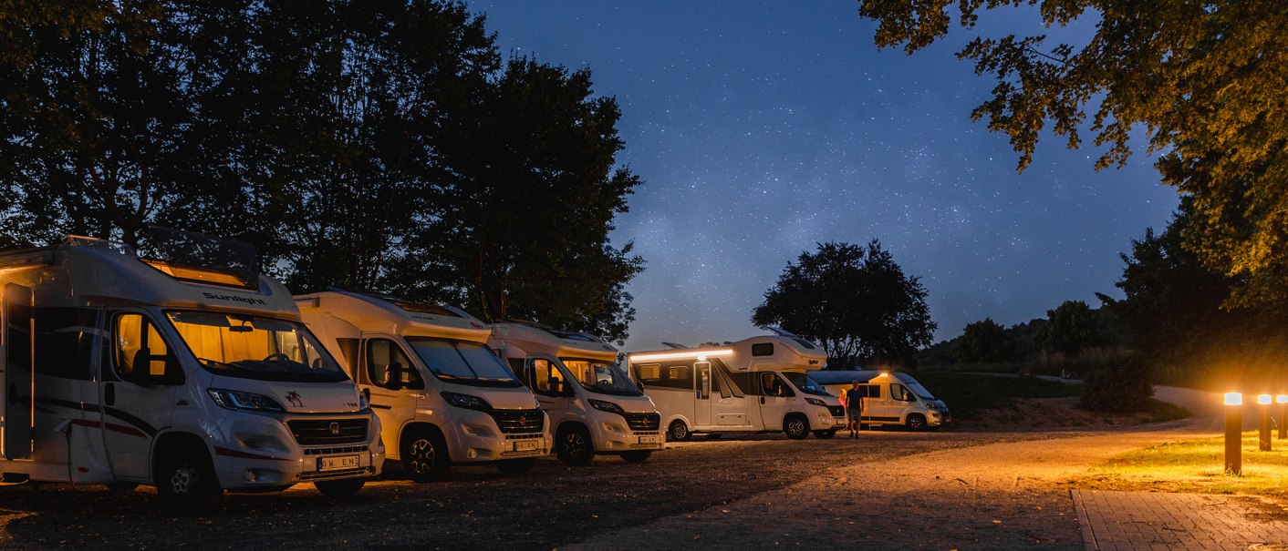 Motorhomes park at night under a starry sky on a pitch surrounded by trees and an illuminated path., © Blu Dolci Artwork