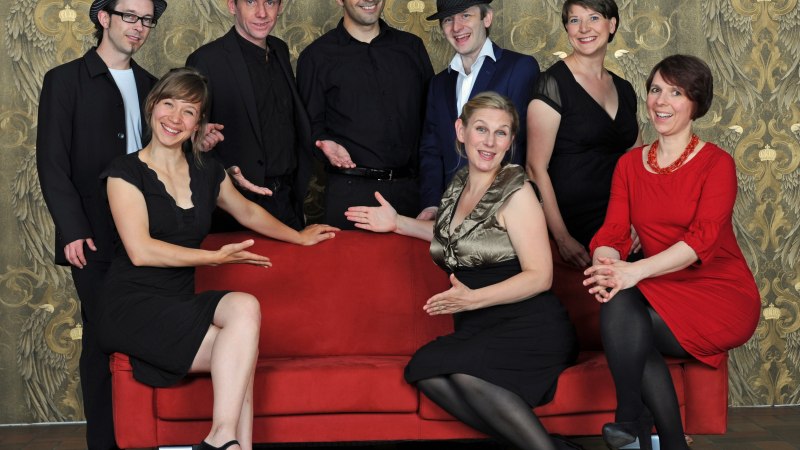 Eight people in elegant clothing pose around a red sofa in front of an artistic background. They smile and point to the sofa., &copy; Yakup Zeyrek