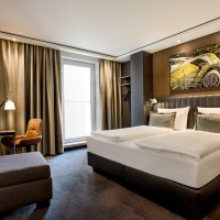 Modernes Hotelzimmer mit Doppelbett, Sofa, Schreibtisch und großem Fenster. An der Wand hängt ein Kunstwerk mit einem Auto-Motiv., © TOMAS Modernes Hotelzimmer mit Doppelbett, Sofa, Schreibtisch und großem Fenster. An der Wand hängt ein Kunstwerk mit einem Auto-Motiv., © TOMAS
