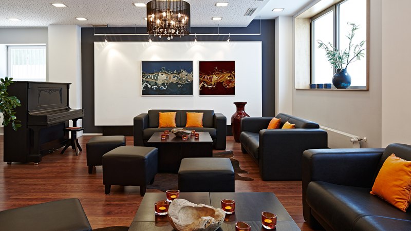 Elegante Lounge mit schwarzen Sofas, orangefarbenen Kissen, Klavier, Kunstwerken und Kerzen auf Holztischen., &copy; Studierendenwerk Stuttgart