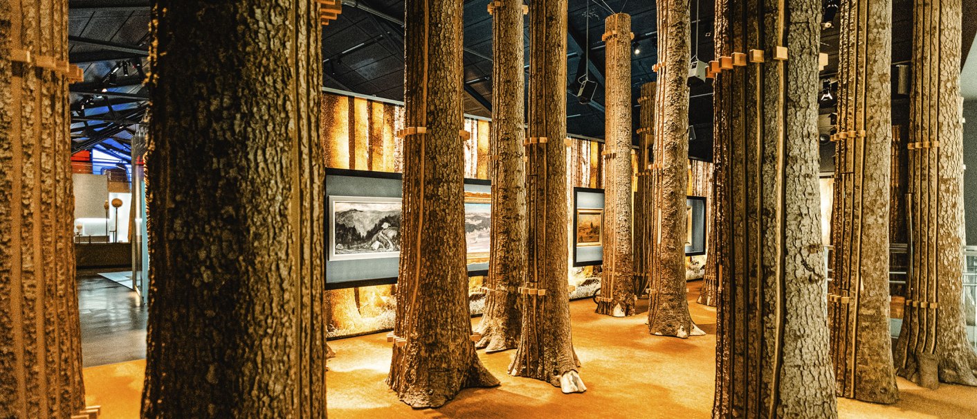 Innenansicht einer Ausstellung mit Baumstämmen als Säulen und Gemälden an den Wänden. Die Beleuchtung ist warm und atmosphärisch., © Stuttgart Marketing GmbH, Sarah Schmid Innenansicht einer Ausstellung mit Baumstämmen als Säulen und Gemälden an den Wänden. Die Beleuchtung ist warm und atmosphärisch., © Stuttgart Marketing GmbH, Sarah Schmid