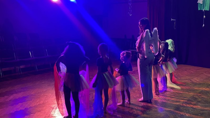 Kinder in bunten T&uuml;llr&ouml;cken und eine Frau mit Engelsfl&uuml;geln proben auf einer B&uuml;hne. Bunte Lichter beleuchten die Szene., &copy; Tathea - Lena Oschowitzer