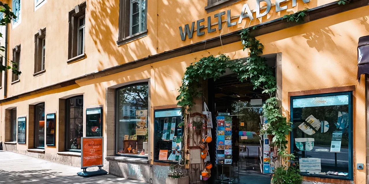 Der Weltladen an einer sonnigen Straßenecke mit Schaufenstern, Pflanzen am Eingang und großem Schriftzug an der Fassade., © Weltladen, Stuttgart Der Weltladen an einer sonnigen Straßenecke mit Schaufenstern, Pflanzen am Eingang und großem Schriftzug an der Fassade., © Weltladen, Stuttgart