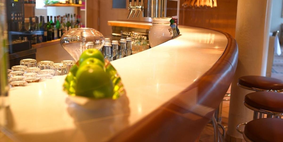 Elegante Bar mit geschwungener Theke, Barhockern und Flaschen im Hintergrund. Im Vordergrund liegen grüne Äpfel auf einem Teller., © Hotel am Park Elegante Bar mit geschwungener Theke, Barhockern und Flaschen im Hintergrund. Im Vordergrund liegen grüne Äpfel auf einem Teller., © Hotel am Park