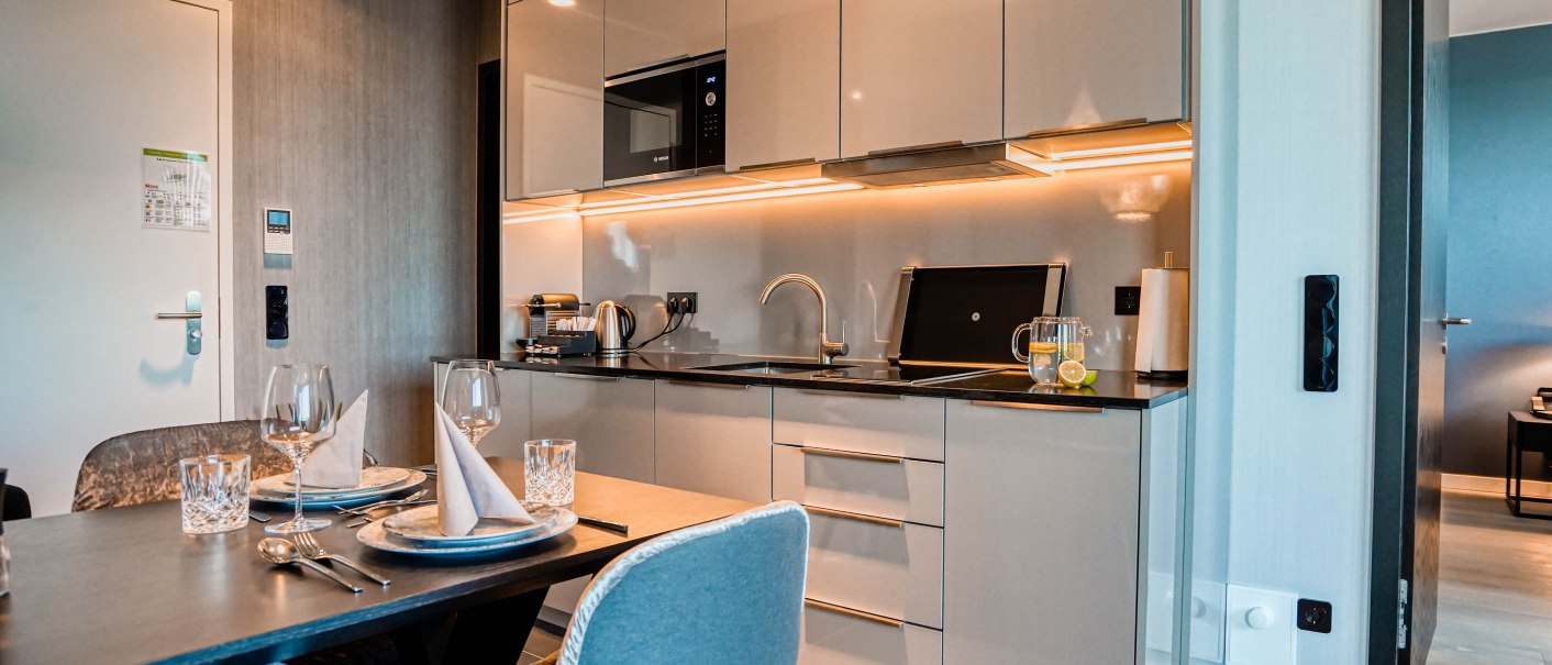 Moderne Küche in einer Suite mit Esstisch, gedeckt für zwei Personen. Helle Schränke, Beleuchtung unter den Schränken, Wasserkocher und Kaffeemaschine vorhanden., © CLOUD N°7 APARTMENTS GmbH Moderne Küche in einer Suite mit Esstisch, gedeckt für zwei Personen. Helle Schränke, Beleuchtung unter den Schränken, Wasserkocher und Kaffeemaschine vorhanden., © CLOUD N°7 APARTMENTS GmbH