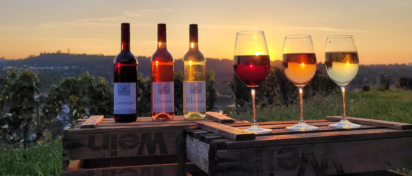 Three wine bottles and three filled glasses stand on wooden crates in the vineyard. In the background, the sun sets over the hills., © Weinbau Glock und Sohn GbR Three wine bottles and three filled glasses stand on wooden crates in the vineyard. In the background, the sun sets over the hills., © Weinbau Glock und Sohn GbR