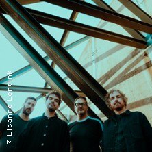 Vier M&auml;nner posieren unter einer modernen Stahl- und Glasstruktur. Die Perspektive ist von unten aufgenommen., &copy; links im Bild