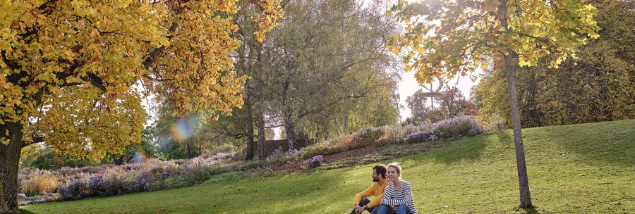 A couple is sitting on a meadow in Rosenstein Park, surrounded by autumnal trees and flowers. The sun is shining through the leaves., &copy; SMG, Christoph D&uuml;pper