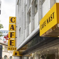 Gelbes Schild des Cafe Nast mit Kaffeetasse-Logo an einem Geb&auml;ude, umgeben von B&auml;umen., &copy; SMG, Sarah Schmid
