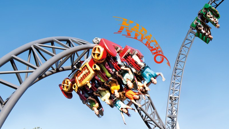 Eine Achterbahn im Erlebnispark Tripsdrill zeigt Menschen in einem Looping unter blauem Himmel. Die Wagen sind rot und grün., © Michael Ehrhart Eine Achterbahn im Erlebnispark Tripsdrill zeigt Menschen in einem Looping unter blauem Himmel. Die Wagen sind rot und grün., © Michael Ehrhart