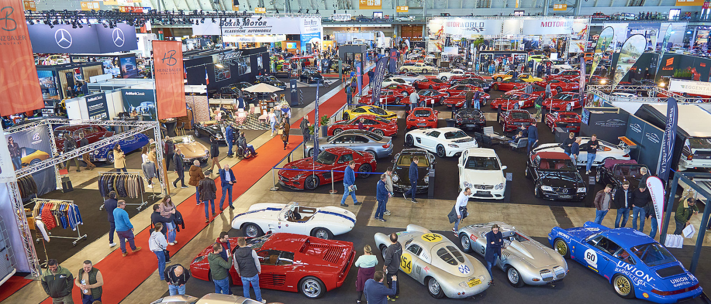 Bird's eye view of a car show with many classic and modern cars, stands and visitors. Brand logos and red carpets are visible., © RETRO CLASSICS® Bird's eye view of a car show with many classic and modern cars, stands and visitors. Brand logos and red carpets are visible., © RETRO CLASSICS®