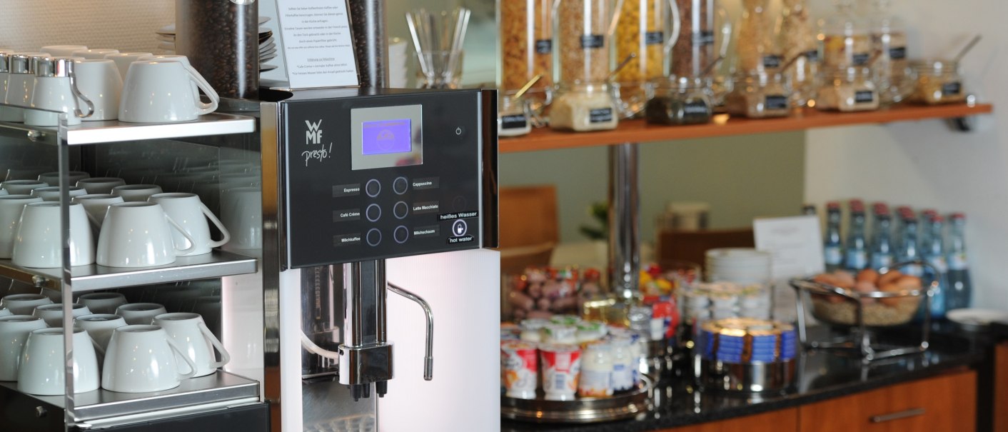 Ein Frühstücksbuffet mit einer Kaffeemaschine, Tassen, Müsli-Spendern, Joghurt und Wasserflaschen auf einer Theke., © TOMAS