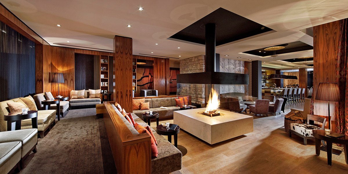 Elegante Lounge mit zentralem Kamin, gem&uuml;tlichen Sofas und einer Bar im Hintergrund. Holzw&auml;nde und warme Beleuchtung schaffen eine einladende Atmosph&auml;re., &copy; Parkhotel Stuttgart Messe-Airport