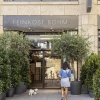 Eingang von Feinkost Böhm, umgeben von grünen Pflanzen. Eine Frau mit einem kleinen weißen Hund betritt das Geschäft., © SMG, Sarah Schmid Eingang von Feinkost Böhm, umgeben von grünen Pflanzen. Eine Frau mit einem kleinen weißen Hund betritt das Geschäft., © SMG, Sarah Schmid