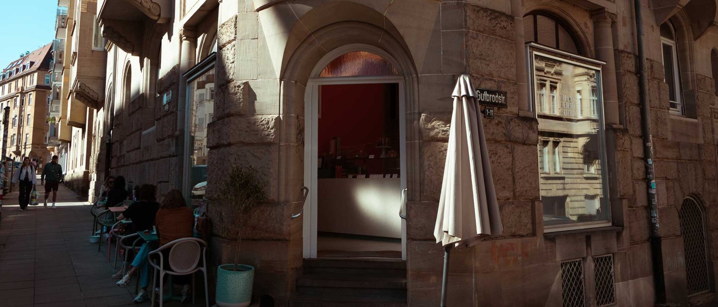 Ein Café in der Gutbrodstraße mit Sitzgelegenheiten im Freien. Menschen sitzen an Tischen, während andere die Straße entlanggehen., © SMG