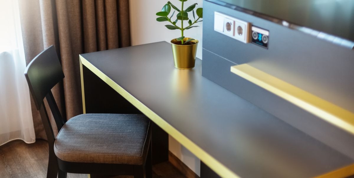 A modern desk with yellow accents, a potted plant, and a chair in front of a window with curtains. Electrical outlets and a Bluetooth connection on the wall., &copy; Novum Hospitality