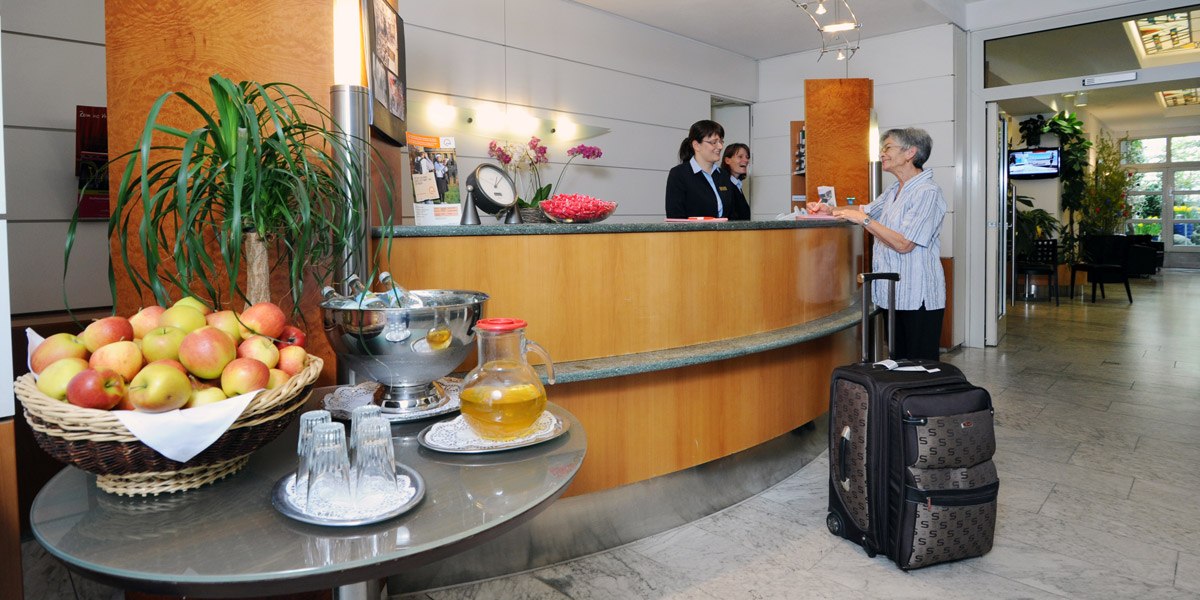 Eine Hotelrezeption mit zwei Mitarbeiterinnen, die einer älteren Frau mit Koffer helfen. Ein Tisch mit Äpfeln und Getränken steht im Vordergrund., © Hotel Unger Eine Hotelrezeption mit zwei Mitarbeiterinnen, die einer älteren Frau mit Koffer helfen. Ein Tisch mit Äpfeln und Getränken steht im Vordergrund., © Hotel Unger
