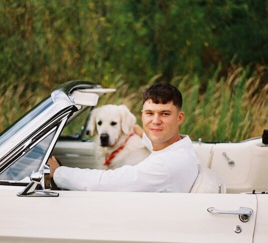 Ein Mann sitzt lächelnd in einem weißen Cabrio, neben ihm ein Hund. Im Hintergrund sind grüne Büsche zu sehen., © Rosenau Kultur e.V.