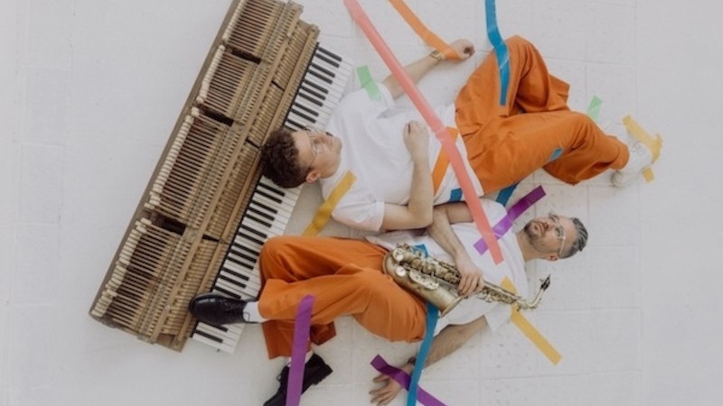 Zwei Männer in orangefarbener Kleidung liegen auf einem weißen Boden, umgeben von bunten Klebebändern. Einer hält ein Saxophon, der andere liegt neben einer Klaviatur., © Renitenztheater Stuttgart e.V. Zwei Männer in orangefarbener Kleidung liegen auf einem weißen Boden, umgeben von bunten Klebebändern. Einer hält ein Saxophon, der andere liegt neben einer Klaviatur., © Renitenztheater Stuttgart e.V.