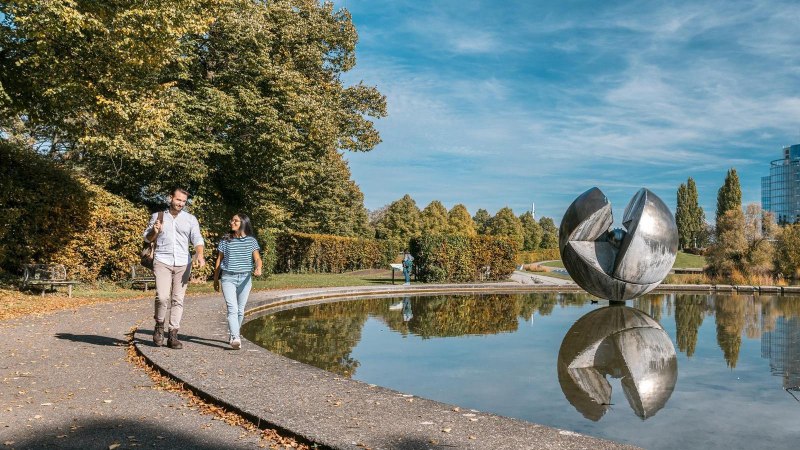 Ein Paar spaziert im Wartbergpark entlang eines Teiches mit einer großen Skulptur und einem modernen Gebäude im Hintergrund., © Stuttgart-Marketing GmbH, Sarah Schmid